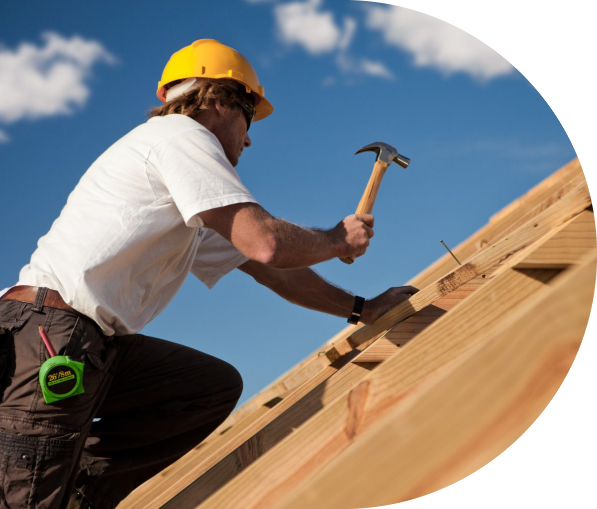 A construction worker in a yellow hard hat hammers a nail into wooden framing under a clear blue sky.